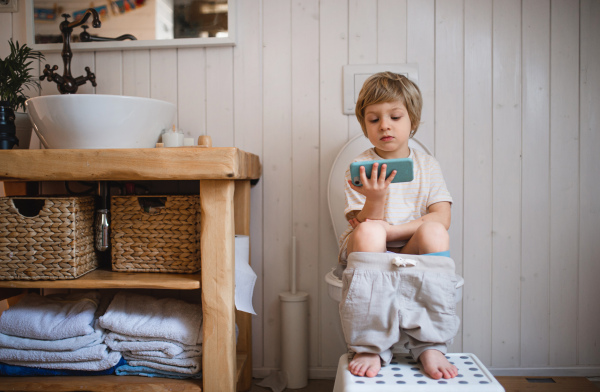 A portrait of cute small boy sitting on toilet indoors at home, using smartphone.