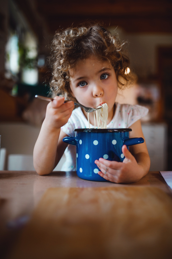 Portrait of sad small girl sitting at table indoors at home, eating spaghetti.