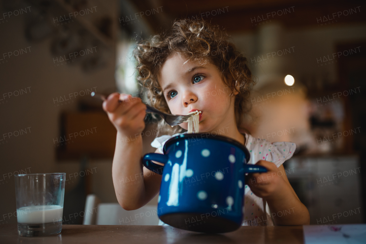 Portrait of sad small girl sitting at table indoors at home, eating spaghetti.