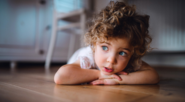 A portrait of cute small girl lying on floor under table indoors at home, looking away.