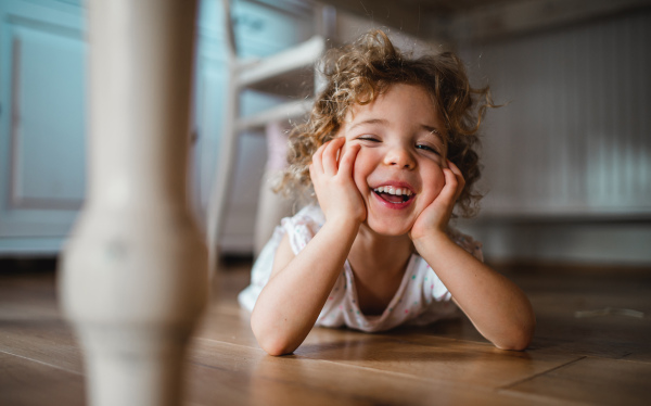 A portrait of cute small girl lying on floor under table indoors at home, looking at camera.