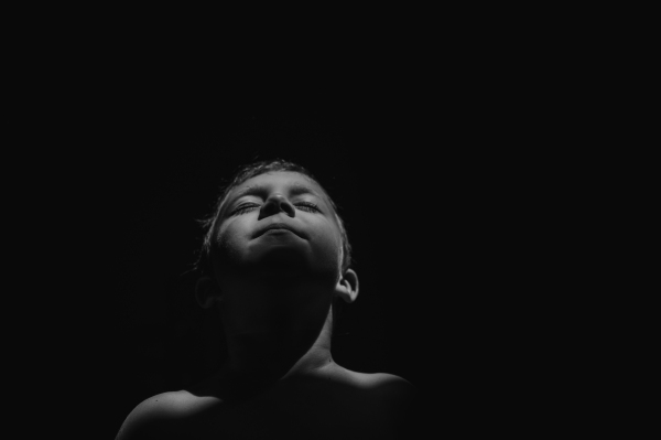 A black and white portrait of little boy with eyes closed raising face on black background