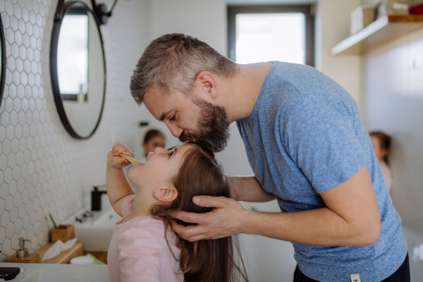 A father kissing his daughter while she is brushing her teeth in bathroom, morning routine concept.