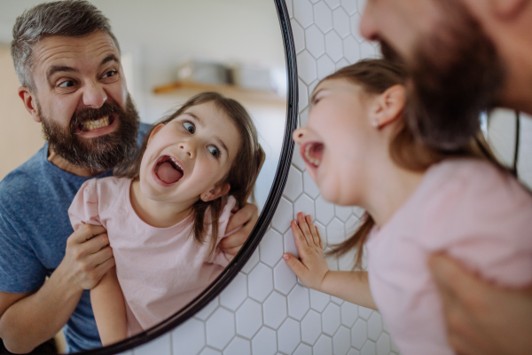 A father having fun with his little daughter, making grimaces in mirror in bathroom at home.