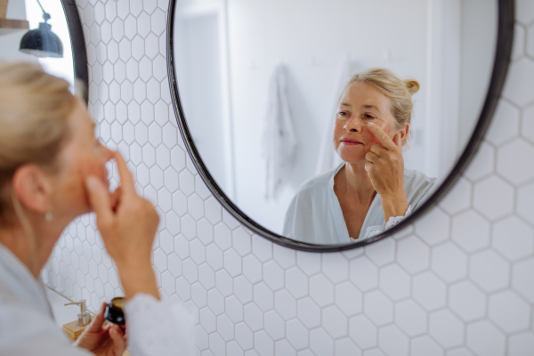 A beautiful senior woman in bathrobe applying natural face cream in bathroom, skin care and morning routine concept.