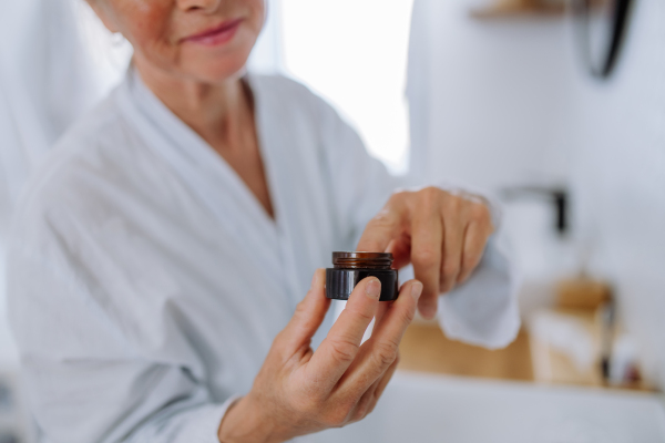 A beautiful senior woman in bathrobe applying natural face cream in bathroom, skin care and morning routine concept.