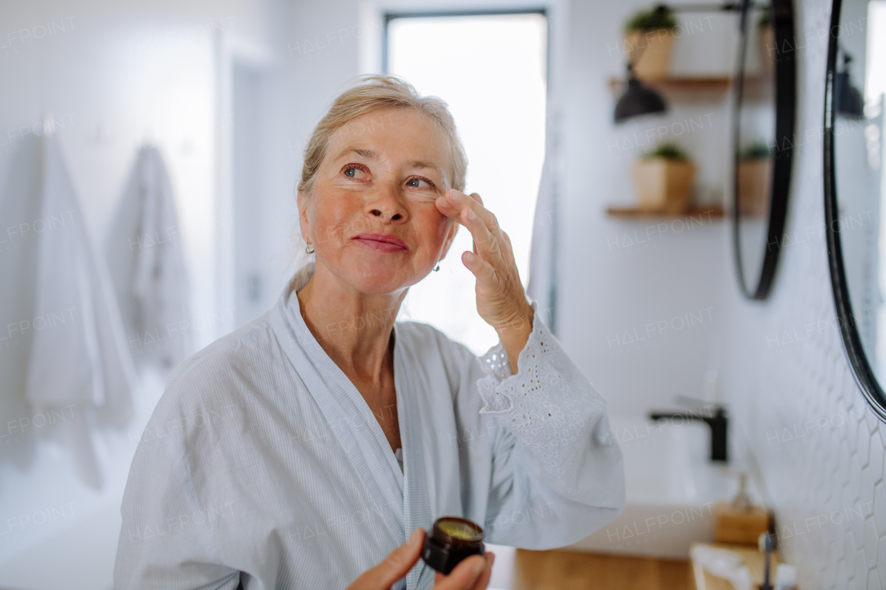 A beautiful senior woman in bathrobe applying natural face cream in bathroom, skin care and morning routine concept.