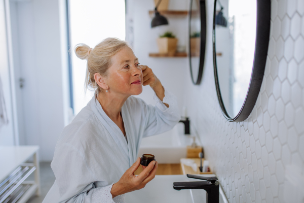 A beautiful senior woman in bathrobe applying natural face cream in bathroom, skin care and morning routine concept.