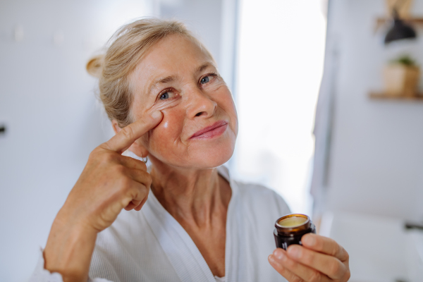 A beautiful senior woman in bathrobe applying natural face cream in bathroom, skin care and morning routine concept.