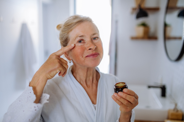 A beautiful senior woman in bathrobe applying natural face cream in bathroom, skin care and morning routine concept.