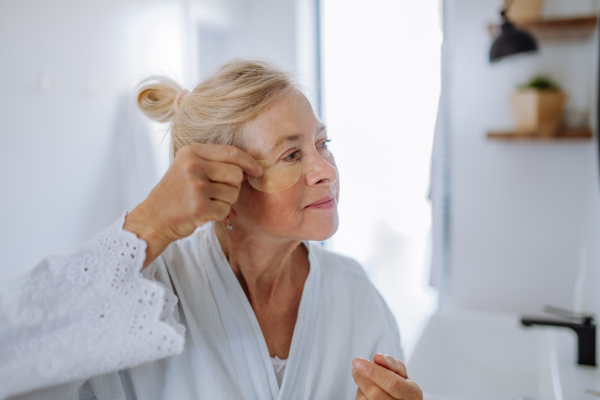 A beautiful senior woman in bathrobe, applying eye patches for puffiness while looking in the mirror