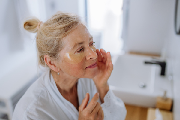 A beautiful senior woman in bathrobe, applying eye patches for puffiness while looking in the mirror