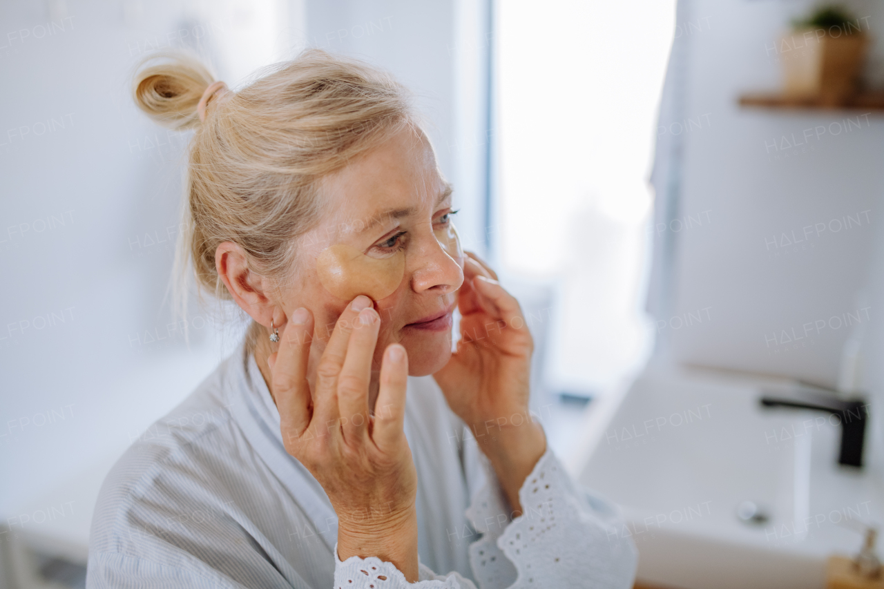 A beautiful senior woman in bathrobe, applying eye patches for puffiness while looking in the mirror