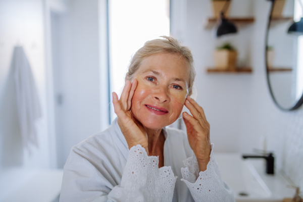 A beautiful senior woman in bathrobe, applying eye patches for puffiness while looking at camera