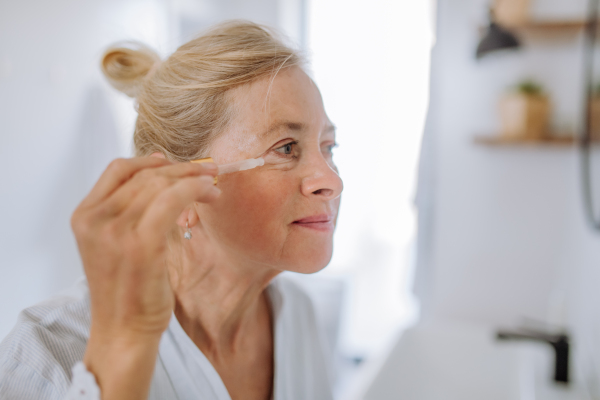 A beautiful senior woman in bathrobe, moisturizing skin by pipette with serum enjoying treatment procedure.