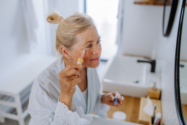 A beautiful senior woman in bathrobe, moisturizing skin by pipette with serum enjoying treatment procedure.