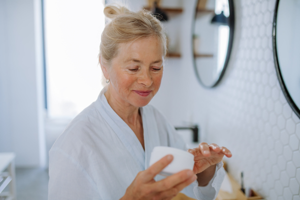 A beautiful senior woman in bathrobe applying natural cream in bathroom, skin care and morning routine concept.