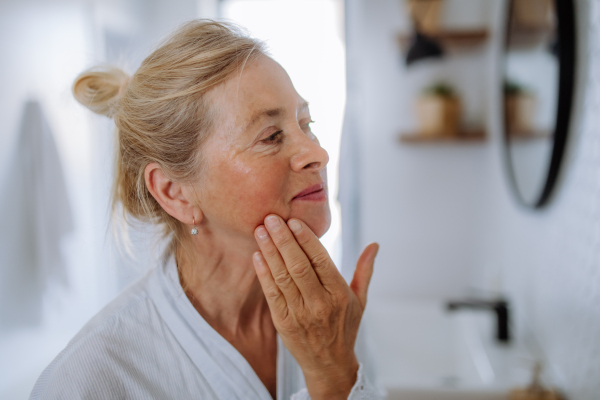 A beautiful senior woman in bathrobe looking at mirror and applying natural face cream in bathroom, skin care concept.