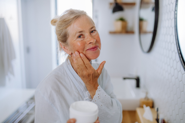 A beautiful senior woman in bathrobe applying natural cream in bathroom, skin care and morning routine concept.