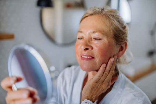 A beautiful senior woman in bathrobe looking at mirror and applying natural face cream in bathroom, skin care concept.