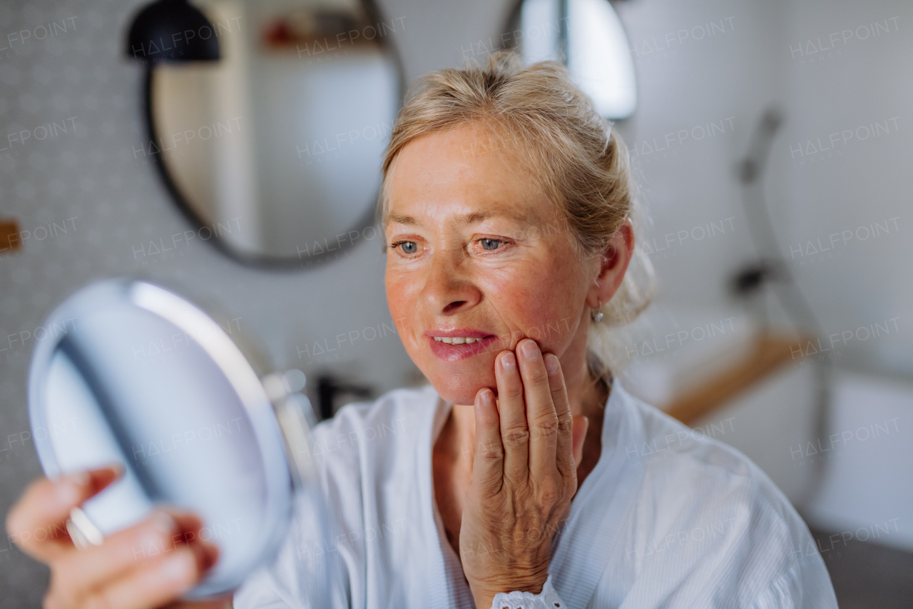 A beautiful senior woman in bathrobe looking at mirror and applying natural face cream in bathroom, skin care concept.