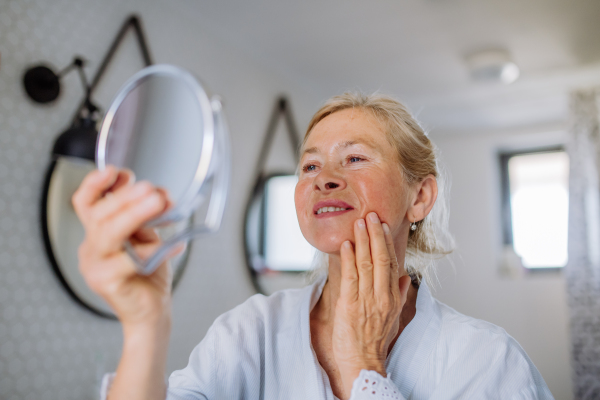 A beautiful senior woman in bathrobe looking at mirror and applying natural face cream in bathroom, skin care concept.