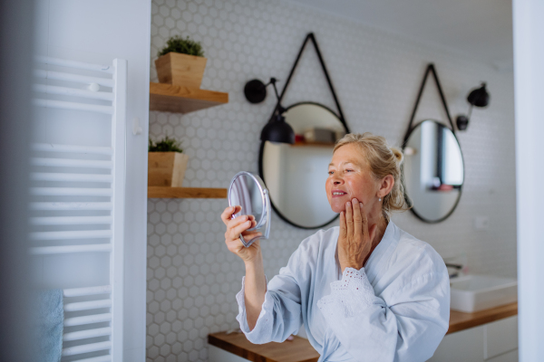 A beautiful senior woman in bathrobe looking at mirror and applying natural face cream in bathroom, skin care concept.