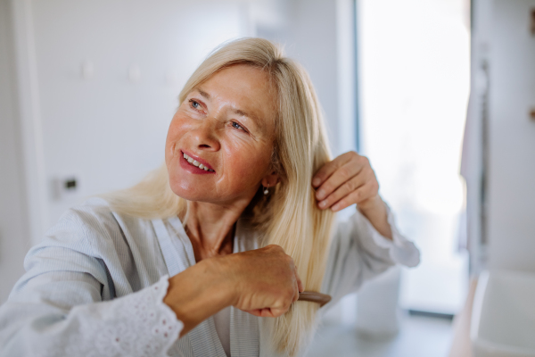 A beautiful senior woman in bathrobe combing hair with wooden comb in bathroom, sustainable lifestyle.