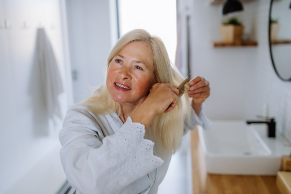 A beautiful senior woman in bathrobe combing hair with wooden comb in bathroom, sustainable lifestyle.