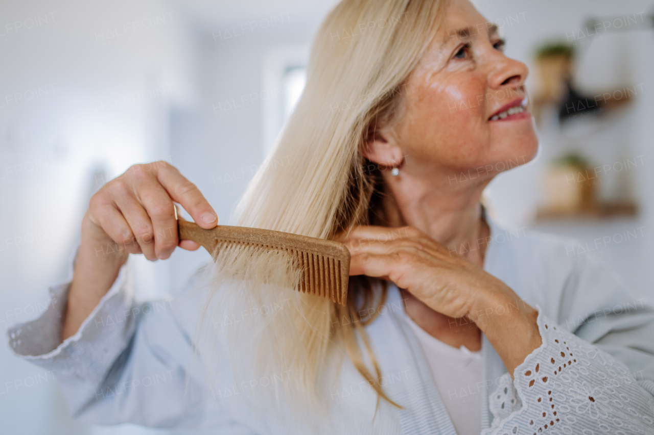 A beautiful senior woman in bathrobe combing hair with wooden comb in bathroom, sustainable lifestyle.