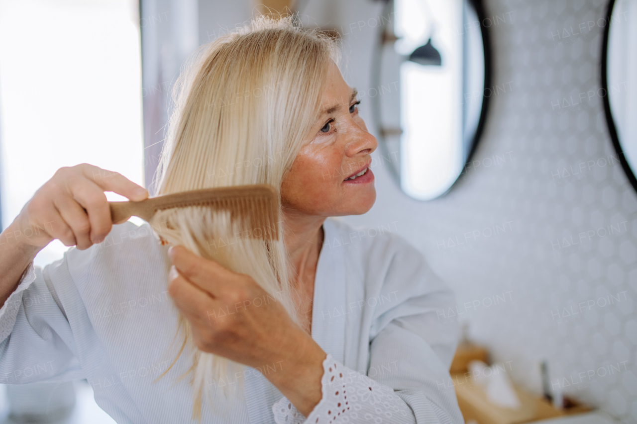 A beautiful senior woman in bathrobe combing hair with wooden comb in bathroom, sustainable lifestyle.