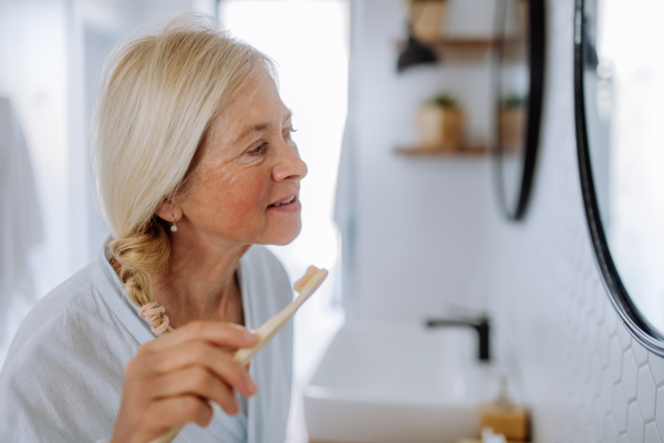 A b eautiful senior woman in bathrobe brushing teeth with eco wooden toothbrush inbathroom, sustainable lifestyle.