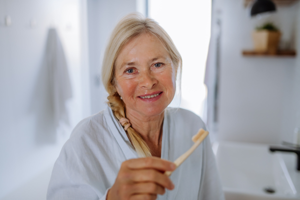 A b eautiful senior woman in bathrobe brushing teeth with eco wooden toothbrush inbathroom, sustainable lifestyle.