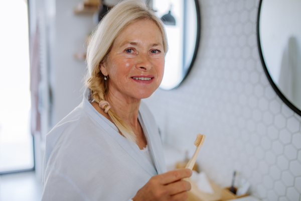 A b eautiful senior woman in bathrobe brushing teeth with eco wooden toothbrush inbathroom, sustainable lifestyle.