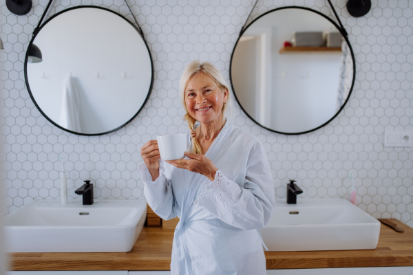 A beautiful senior woman in bathrobe drinking tea in bathroom, relax and wellness concept.