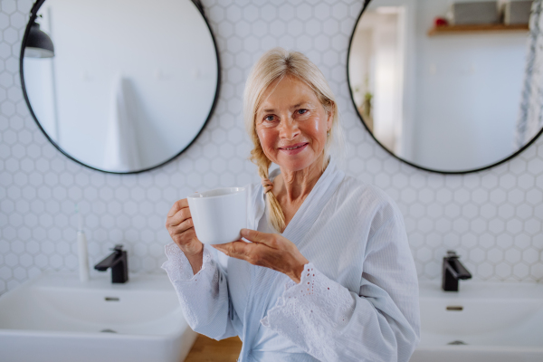 A beautiful senior woman in bathrobe drinking tea in bathroom, relax and wellness concept.