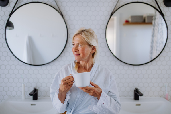 A beautiful senior woman in bathrobe drinking tea in bathroom, relax and wellness concept.