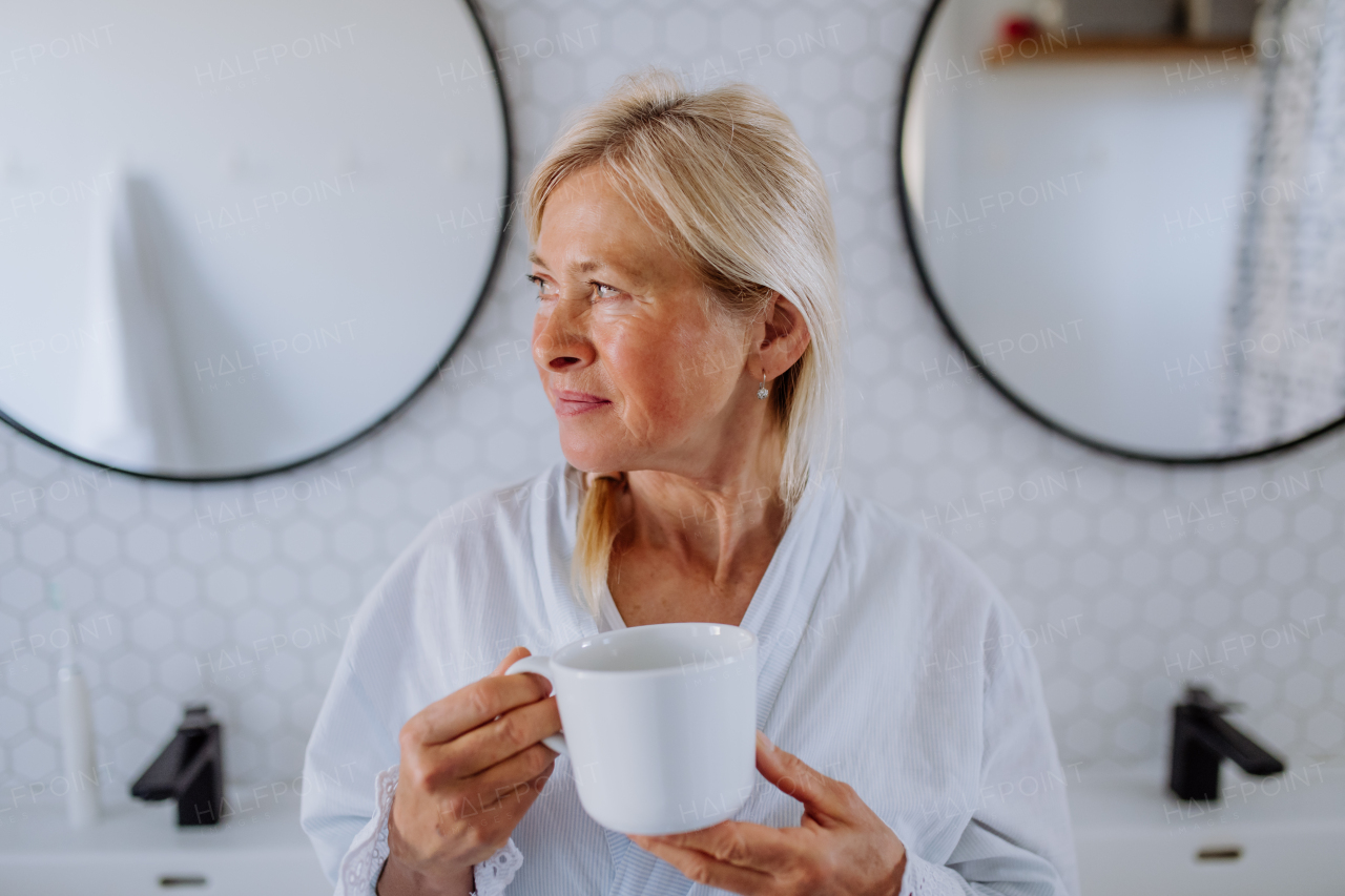 A beautiful senior woman in bathrobe drinking tea in bathroom, relax and wellness concept.