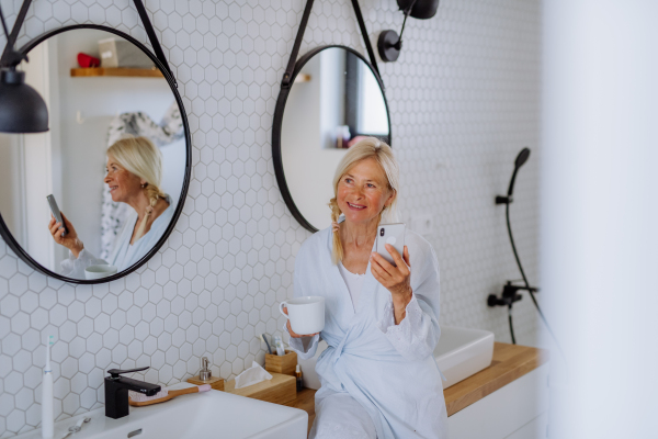 A beautiful senior woman in bathrobe drinking tea and using smartphone in bathroom, relax and wellness concept.