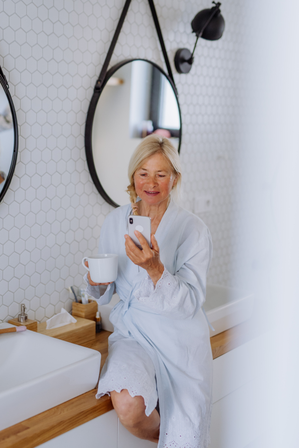 A beautiful senior woman in bathrobe drinking tea and using smartphone in bathroom, relax and wellness concept.