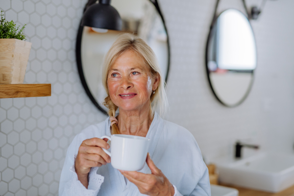 A beautiful senior woman in bathrobe drinking tea in bathroom, relax and wellness concept.