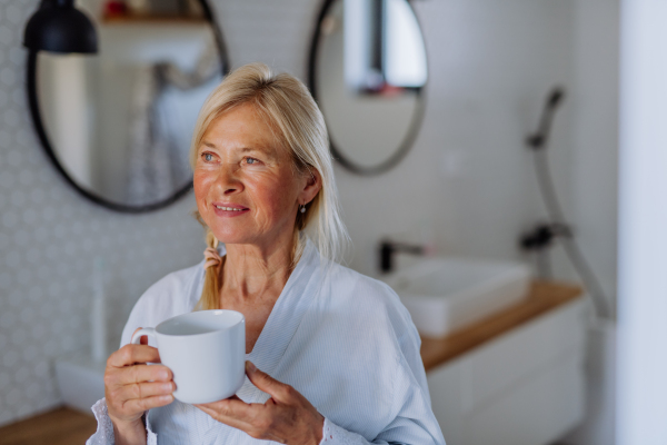 A beautiful senior woman in bathrobe drinking tea in bathroom, relax and wellness concept.