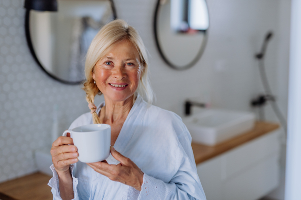 A beautiful senior woman in bathrobe drinking tea in bathroom, relax and wellness concept.