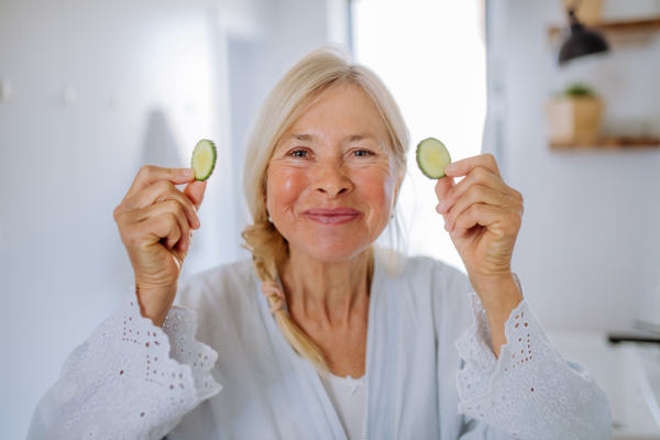 A beautiful senior woman in bathrobe applying cucumber face mask in bathroom, skin care concept.