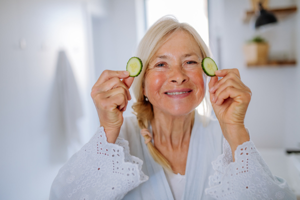 A beautiful senior woman in bathrobe applying cucumber face mask in bathroom, skin care concept.