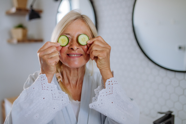 A beautiful senior woman in bathrobe applying cucumber face mask in bathroom, skin care concept.