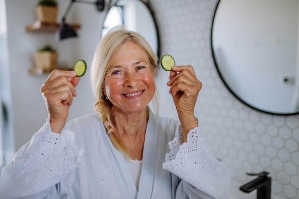 A beautiful senior woman in bathrobe applying cucumber face mask in bathroom, skin care concept.