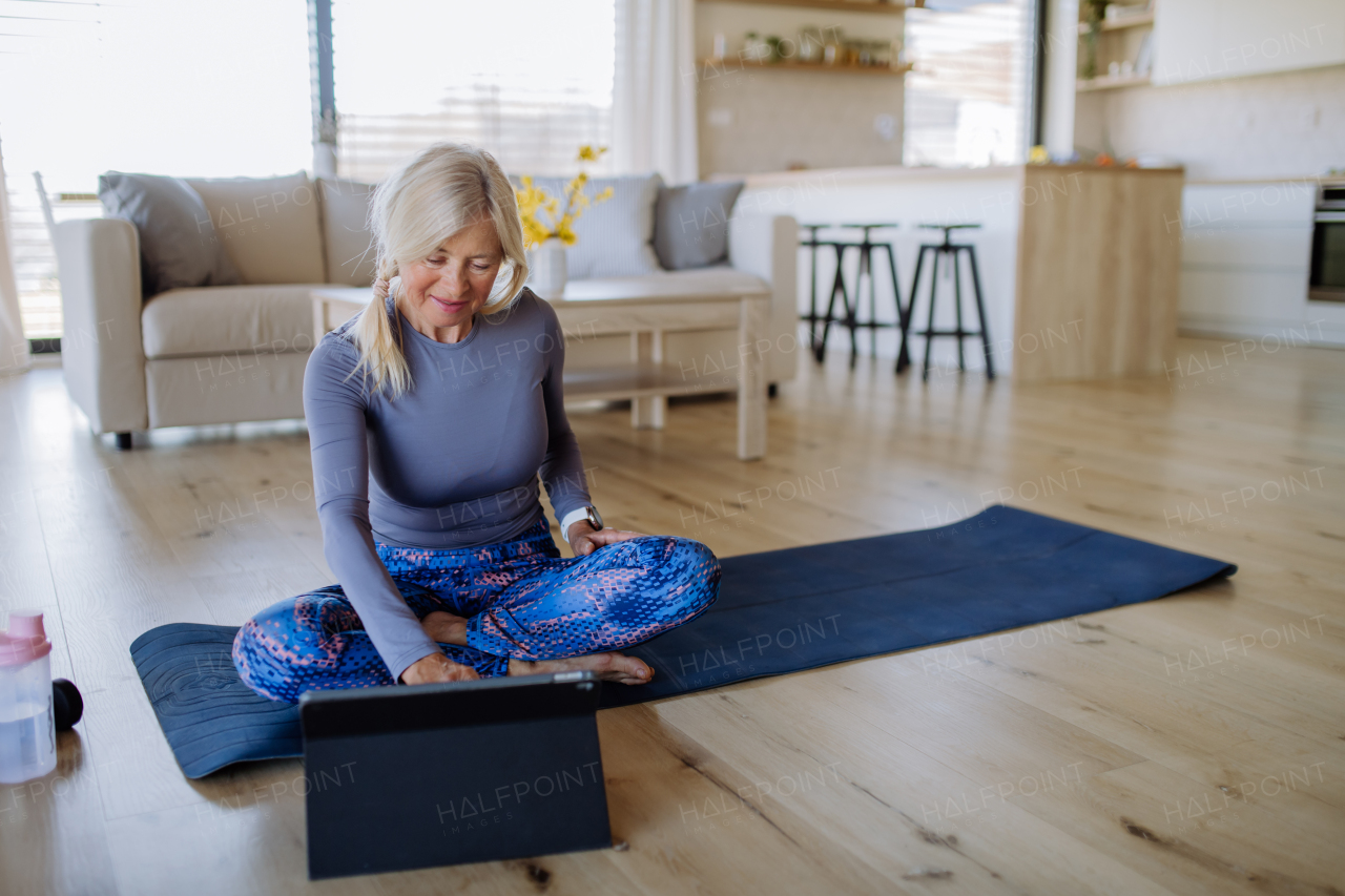 A fit senior woman doing stretching exercise at home with tutorial on tablet, active lifestyle concept.