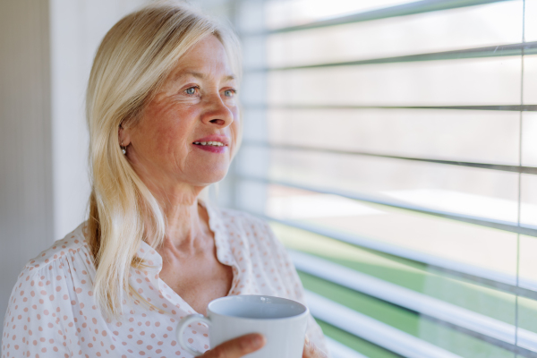 An attractive senior woman drinking tea and standing by window at home.