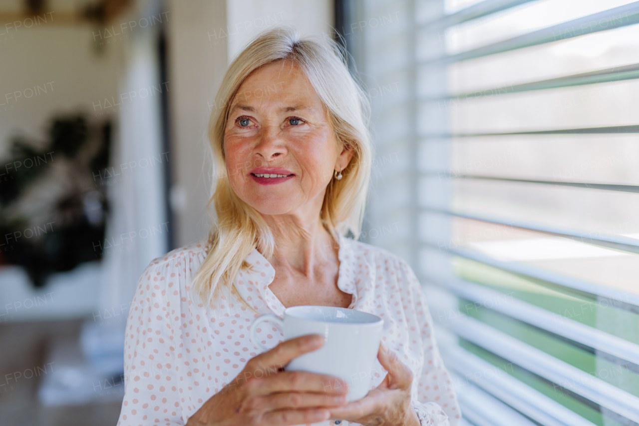 An attractive senior woman drinking tea and standing by window at home.